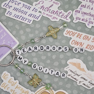 May include: A keychain with the words "TEARDROPS ON MY GUITAR" spelled out in white letter beads. It features green beads, butterfly charms, and a silver keyring. Surrounding the keychain are stickers with song lyrics.