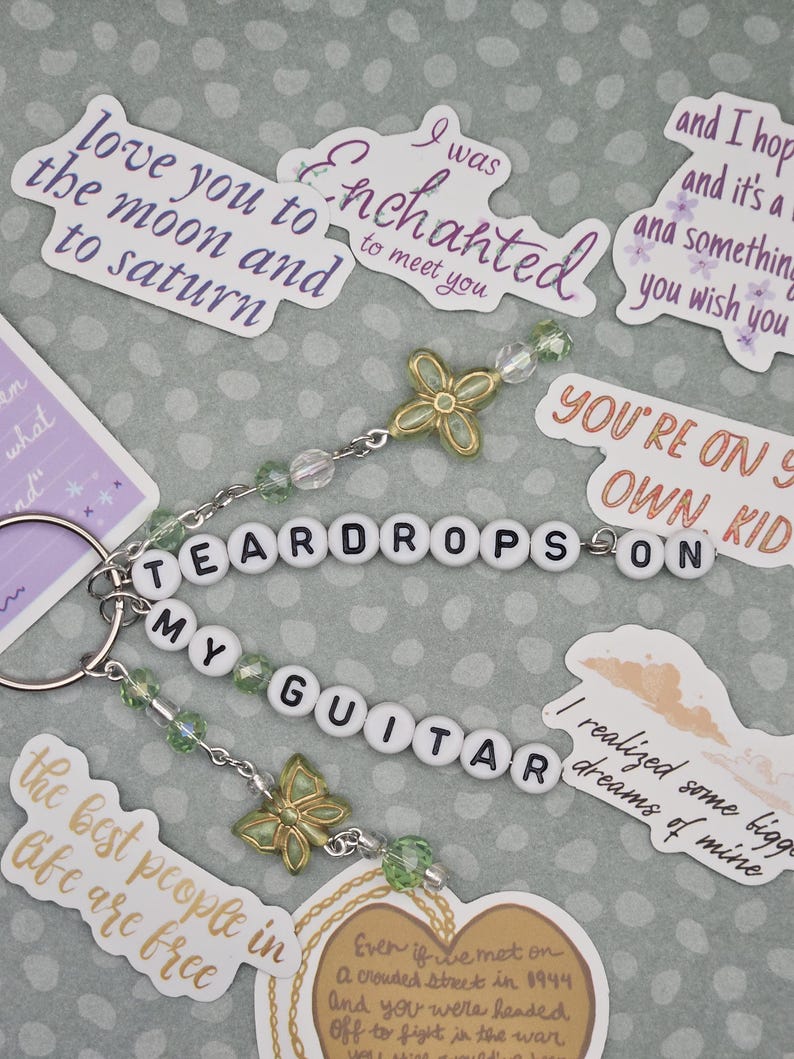 May include: A keychain with the words "TEARDROPS ON MY GUITAR" spelled out in white letter beads. It features green beads, butterfly charms, and a silver keyring. Surrounding the keychain are stickers with song lyrics.
