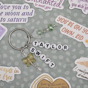 May include: A silver keychain with the name "TAYLOR SWIFT" spelled out in white letter beads. It features a gold butterfly charm and dangling beads. Surrounding the keychain are stickers with song lyrics and phrases.