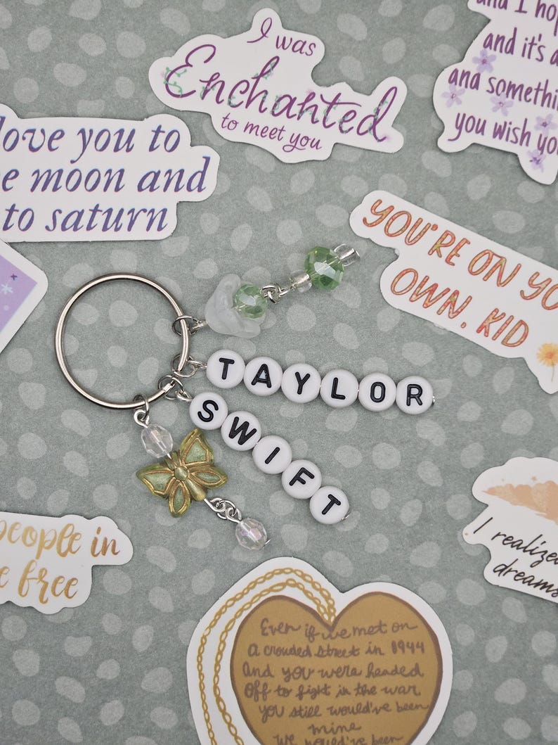 May include: A silver keychain with the name "TAYLOR SWIFT" spelled out in white letter beads. It features a gold butterfly charm and dangling beads. Surrounding the keychain are stickers with song lyrics and phrases.