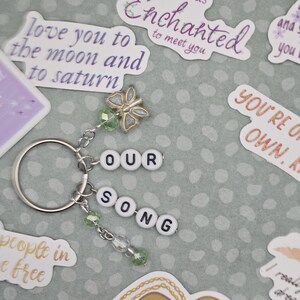 May include: A silver keychain with the words "OUR SONG" spelled out in white letter beads. It features a butterfly charm and green beaded accents. Surrounding the keychain are various stickers with romantic phrases.