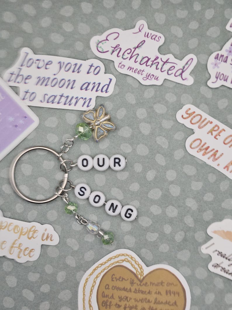 May include: A silver keychain with the words "OUR SONG" spelled out in white letter beads. It features a butterfly charm and green beaded accents. Surrounding the keychain are various stickers with romantic phrases.