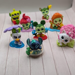 Puede incluir: Una colección de pequeñas figuras coloridas que representan varios personajes de dibujos animados. Las figuras incluyen un extraterrestre verde, un personaje pelirrojo, Mickey Mouse y otros, cada uno con diseños únicos. Están dispuestas sobre una superficie de madera clara.