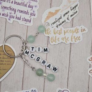 May include: A silver keychain with the name "TIM MCGRAW" spelled out in white letter beads. The keychain features green and clear beads, and is surrounded by decorative stickers with inspirational quotes.