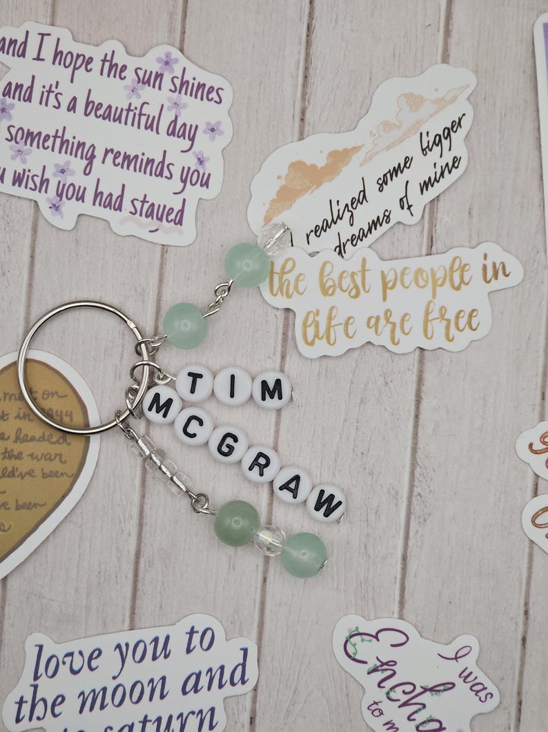 May include: A silver keychain with the name "TIM MCGRAW" spelled out in white letter beads. The keychain features green and clear beads, and is surrounded by decorative stickers with inspirational quotes.