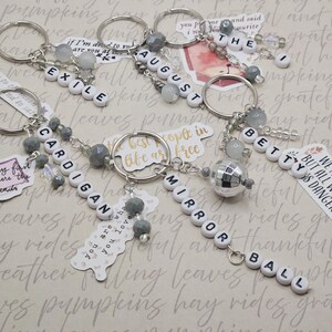 May include: Keyrings with white letter beads spelling out words including "EXILE", "AUGUST", "CARDIGAN", "MIRROR BALL", and "BETTY". Each keyring features silver-tone hardware, gray beads, and a decorative charm. The background has a patterned design.