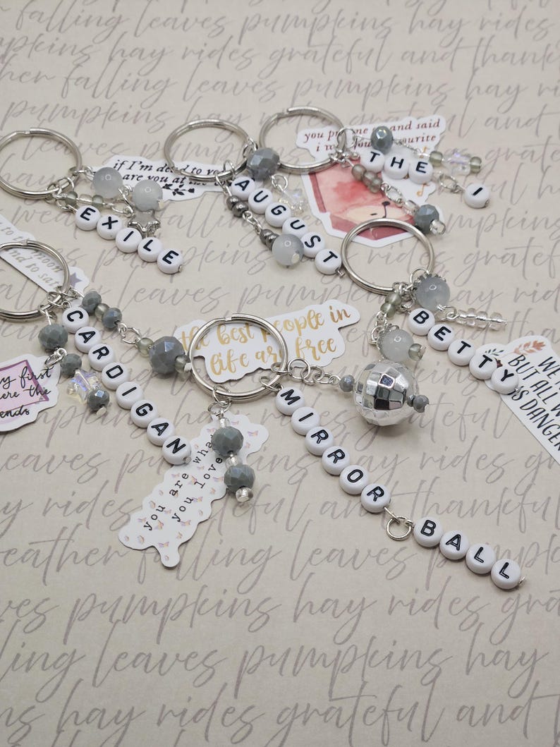 May include: Keyrings with white letter beads spelling out words including "EXILE", "AUGUST", "CARDIGAN", "MIRROR BALL", and "BETTY". Each keyring features silver-tone hardware, gray beads, and a decorative charm. The background has a patterned design.