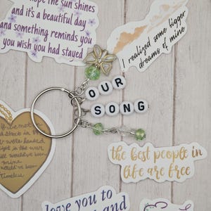 May include: A silver keychain with the words "OUR SONG" spelled out in white letter beads. The keychain is adorned with green beads and a decorative butterfly charm. Surrounding the keychain are various stickers with quotes.