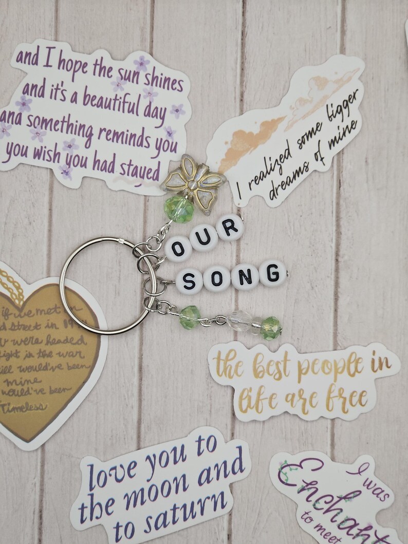 May include: A silver keychain with the words "OUR SONG" spelled out in white letter beads. The keychain is adorned with green beads and a decorative butterfly charm. Surrounding the keychain are various stickers with quotes.