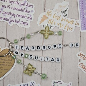 May include: A keychain with the words "TEARDROPS ON MY GUITAR" spelled out in white letter beads. The keychain features green beads, gold-colored butterfly charms, and a gold-colored flower charm. Decorative stickers with quotes surround the keychain.