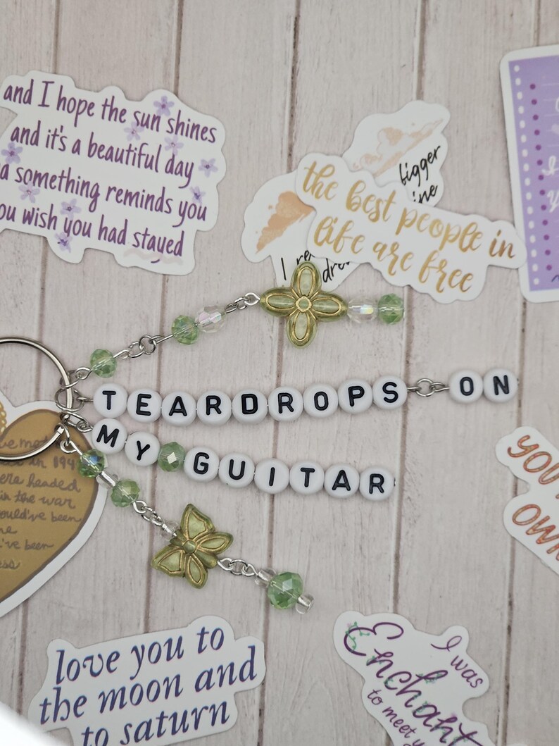 May include: A keychain with the words "TEARDROPS ON MY GUITAR" spelled out in white letter beads. The keychain features green beads, gold-colored butterfly charms, and a gold-colored flower charm. Decorative stickers with quotes surround the keychain.