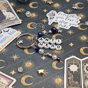 May include: A silver keyring spelling "ANTI HERO" in white letter beads, with blue and clear bead accents, star charms, and a crescent moon charm. Tarot cards and a 12-sided die are visible in the background, on a patterned surface.