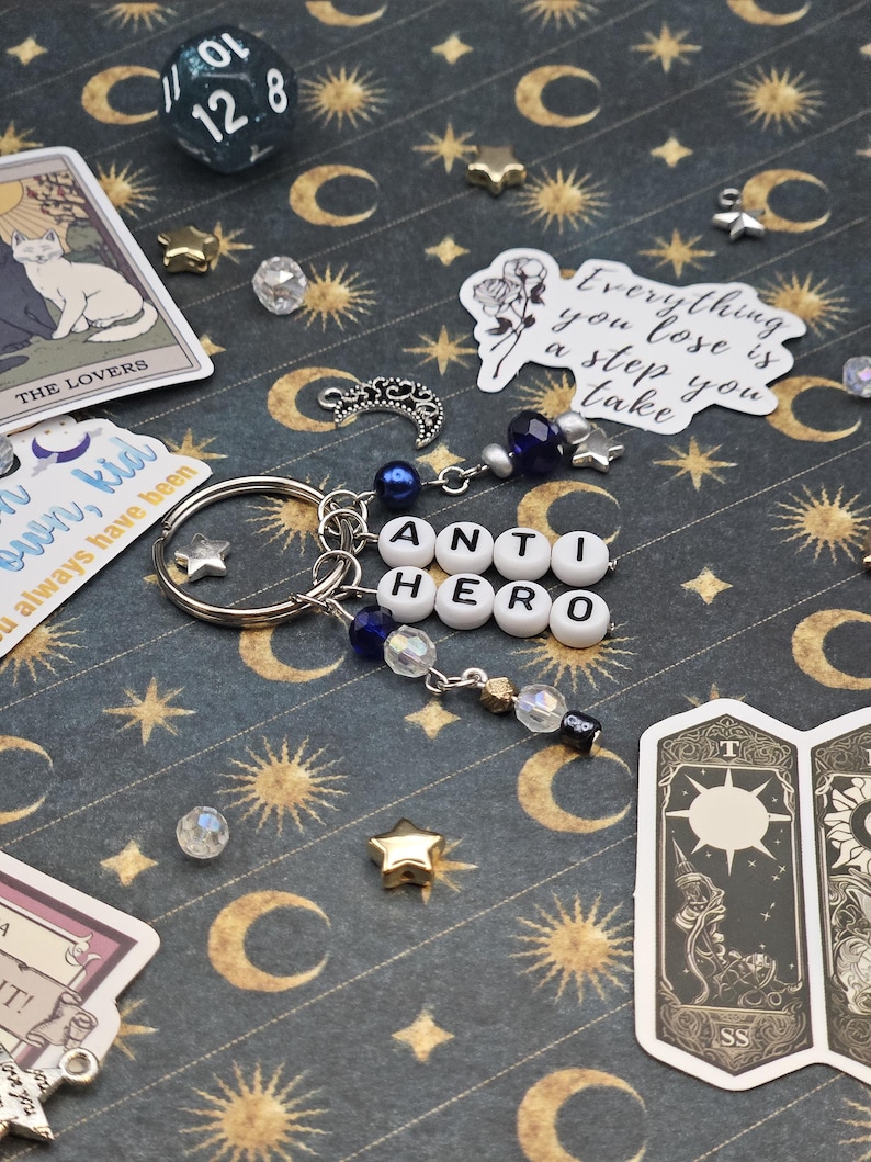May include: A silver keyring spelling "ANTI HERO" in white letter beads, with blue and clear bead accents, star charms, and a crescent moon charm. Tarot cards and a 12-sided die are visible in the background, on a patterned surface.