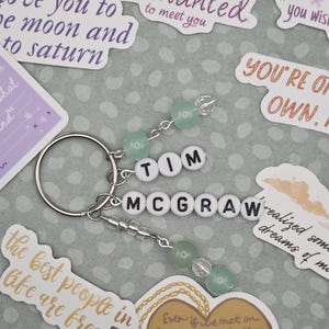 May include: A silver keychain with the name "TIM MCGRAW" spelled out in white letter beads. The keychain features green and clear beads and is surrounded by stickers with phrases like "I was Enchanted to meet you."