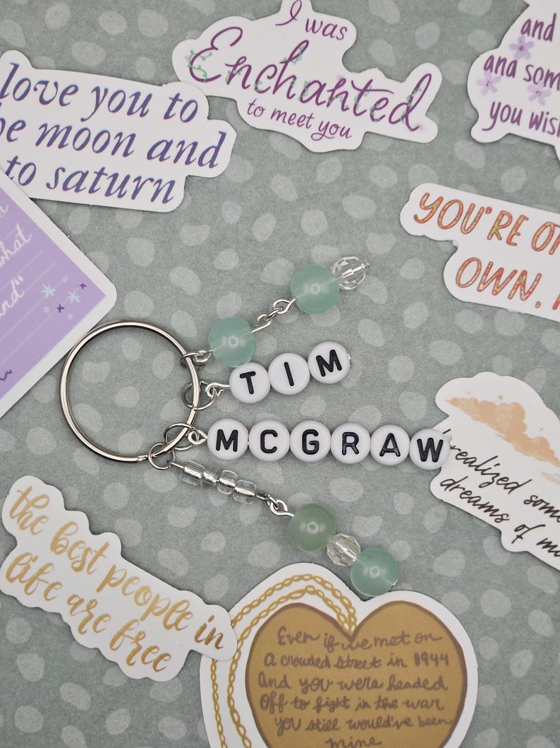 May include: A silver keychain with the name "TIM MCGRAW" spelled out in white letter beads. The keychain features green and clear beads and is surrounded by stickers with phrases like "I was Enchanted to meet you."