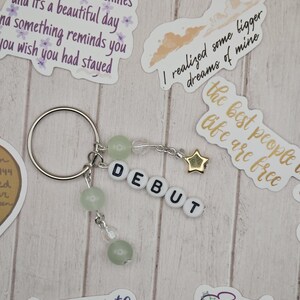 May include: A silver keychain with the word "DEBUT" spelled out in white letter beads. The keychain features green beads and a gold star charm. Decorative stickers with text are scattered around the keychain.