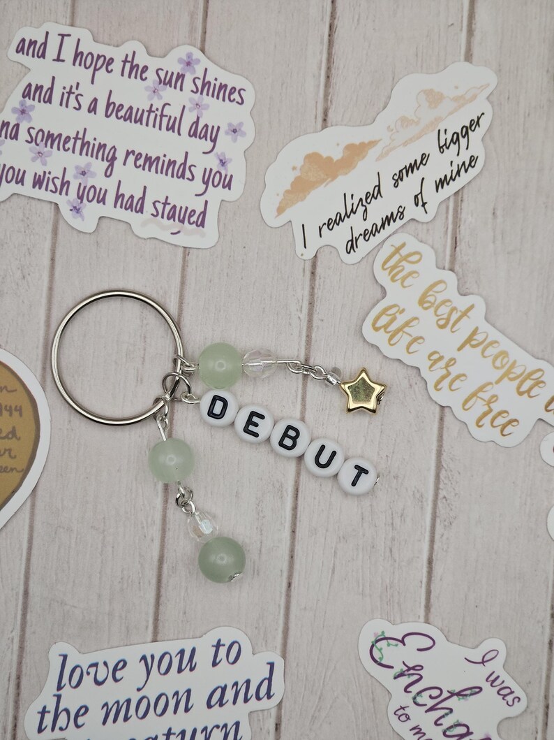 May include: A silver keychain with the word "DEBUT" spelled out in white letter beads. The keychain features green beads and a gold star charm. Decorative stickers with text are scattered around the keychain.