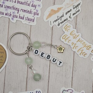 May include: A silver keychain with the word "DEBUT" spelled out in white letter beads. The keychain features green beads and a gold star charm. Decorative stickers with text are scattered around the keychain.