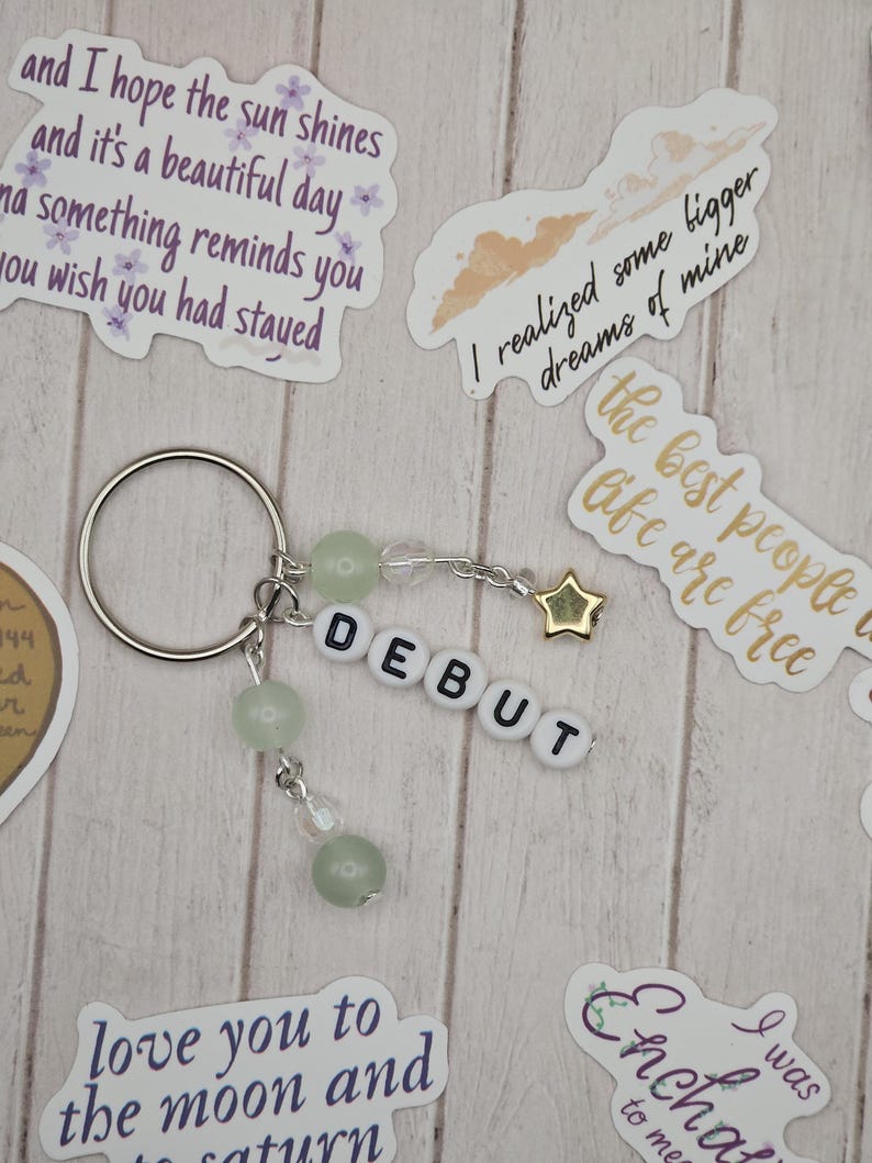 May include: A silver keychain with the word "DEBUT" spelled out in white letter beads. The keychain features green beads and a gold star charm. Decorative stickers with text are scattered around the keychain.
