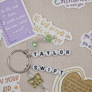 May include: A keychain with the name "TAYLOR SWIFT" spelled out in white letter beads. It includes a gold butterfly charm and clear crystal accents. The background features stickers with song lyrics and quotes.