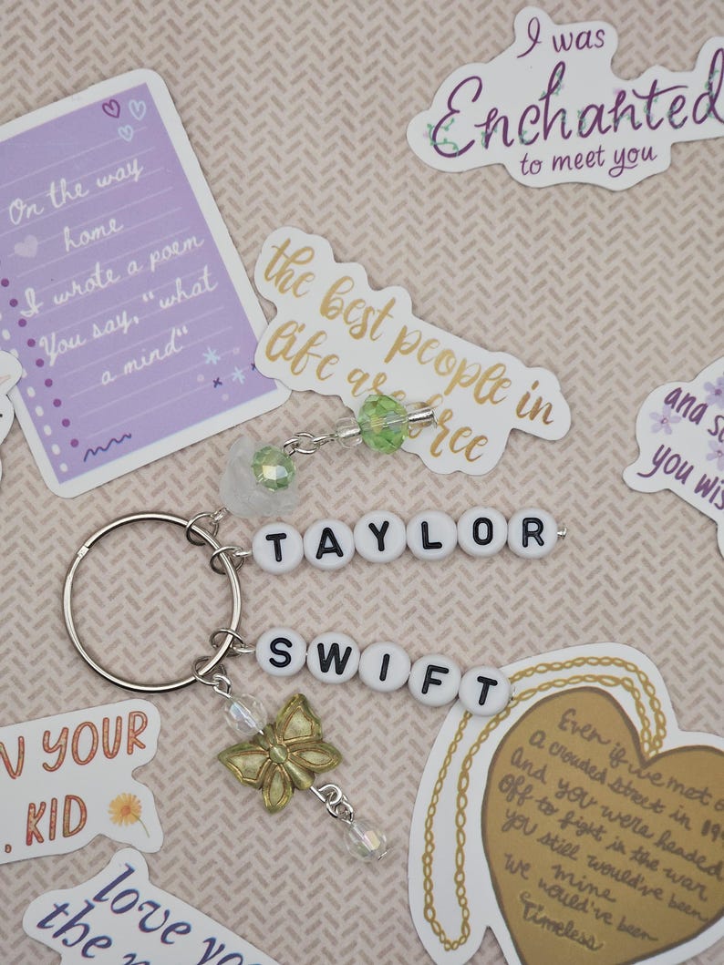 May include: A keychain with the name "TAYLOR SWIFT" spelled out in white letter beads. It includes a gold butterfly charm and clear crystal accents. The background features stickers with song lyrics and quotes.