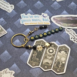 May include: A gold-coloured keyring with a beaded detail and the word "MIDNIGHT" spelled out in green letter beads. A tarot card-style charm with three panels is attached. The background features stickers with phrases like "You're on your own, kid."