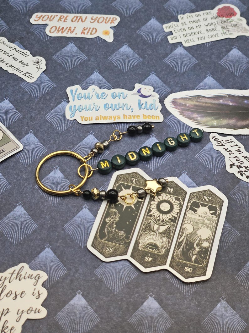 May include: A gold-coloured keyring with a beaded detail and the word "MIDNIGHT" spelled out in green letter beads. A tarot card-style charm with three panels is attached. The background features stickers with phrases like "You're on your own, kid."