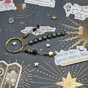 May include: A handmade keychain spelling "MIDNIGHT" in green and gold beads. It has a gold ring, star charms, and decorative beads. The background is dark with gold star and sunburst designs. Includes text stickers.