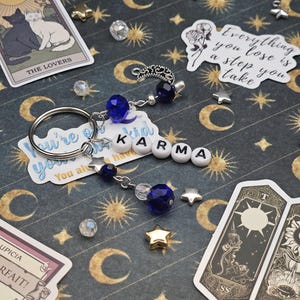 May include: A keychain spelling "KARMA" with white letter beads, blue crystal beads, and a silver charm. The background is a dark blue surface with gold moons, stars, and suns, and tarot cards. The image also includes a 12-sided die.