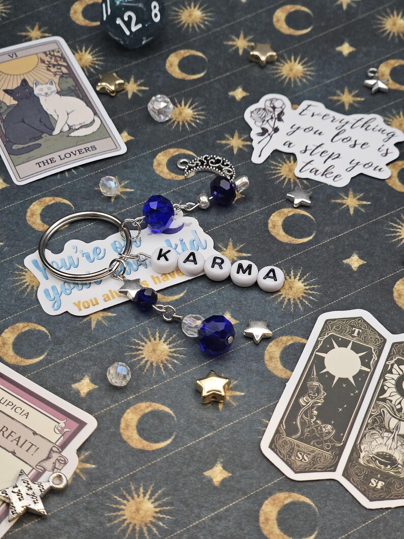 May include: A keychain spelling "KARMA" with white letter beads, blue crystal beads, and a silver charm. The background is a dark blue surface with gold moons, stars, and suns, and tarot cards. The image also includes a 12-sided die.