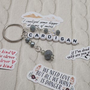 May include: A silver keyring with the word "CARDIGAN" spelled out in white letter beads. The keyring has grey beaded accents and is surrounded by stickers with phrases like "We need love but all we want is danger."