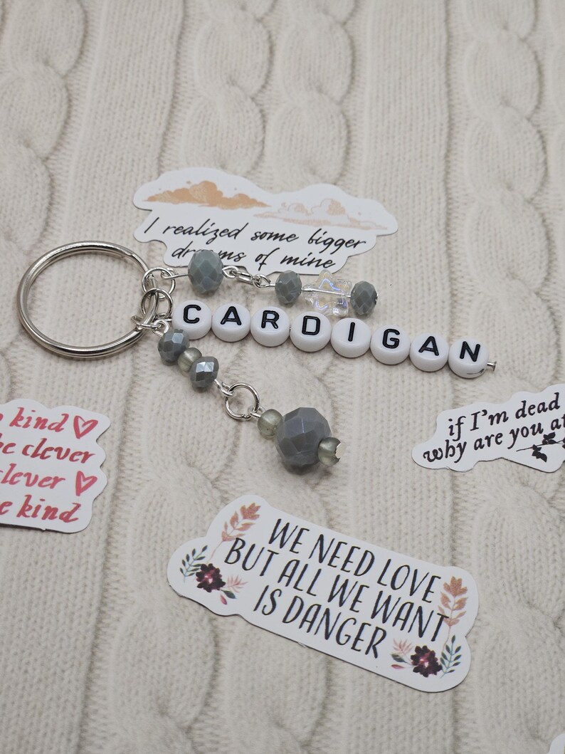 May include: A silver keyring with the word "CARDIGAN" spelled out in white letter beads. The keyring has grey beaded accents and is surrounded by stickers with phrases like "We need love but all we want is danger."