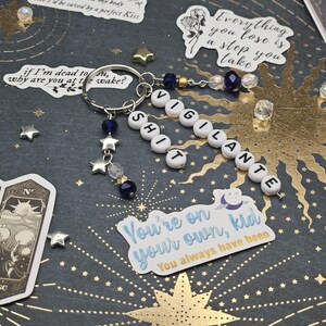 May include: A keychain spelling "VIGILANTE SHIT" with white letter beads, blue and clear beads, and star charms. The keyring is silver. The dark background has star and sunburst patterns, plus stickers with text.