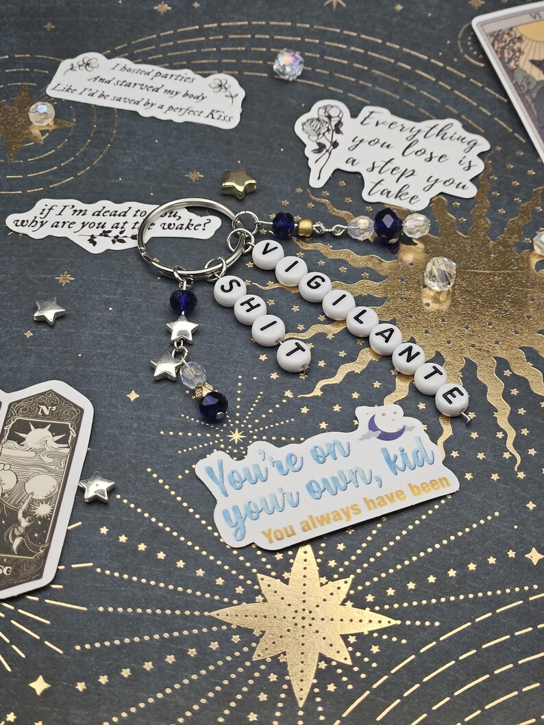 May include: A keychain spelling "VIGILANTE SHIT" with white letter beads, blue and clear beads, and star charms. The keyring is silver. The dark background has star and sunburst patterns, plus stickers with text.