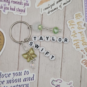 May include: A keychain with the name "TAYLOR SWIFT" spelled out in white letter beads. It features a silver butterfly charm, green and clear beads, and a silver keyring. Decorative stickers with text are scattered around the keychain.