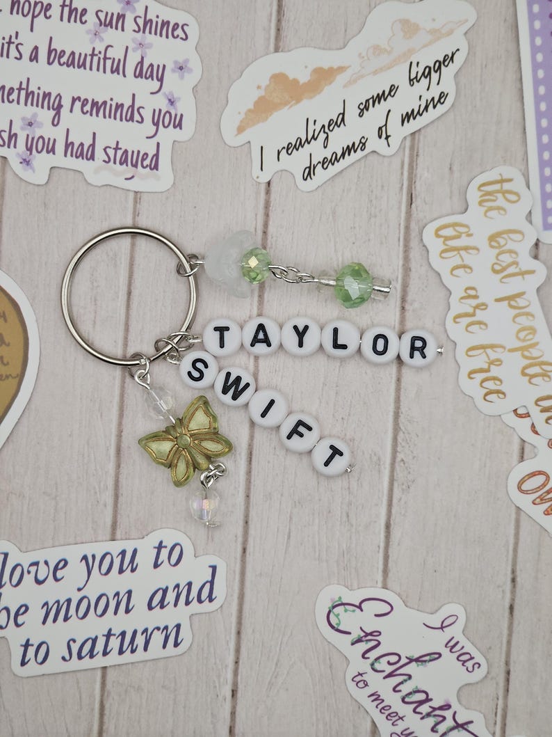 May include: A keychain with the name "TAYLOR SWIFT" spelled out in white letter beads. It features a silver butterfly charm, green and clear beads, and a silver keyring. Decorative stickers with text are scattered around the keychain.