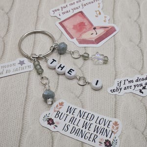 May include: A silver keychain with the word "THE I" spelled out in white letter beads. The keychain has decorative gray beads and charms. Stickers with text and illustrations, including "WE NEED LOVE BUT ALL WE WANT IS DANGER", are scattered around.
