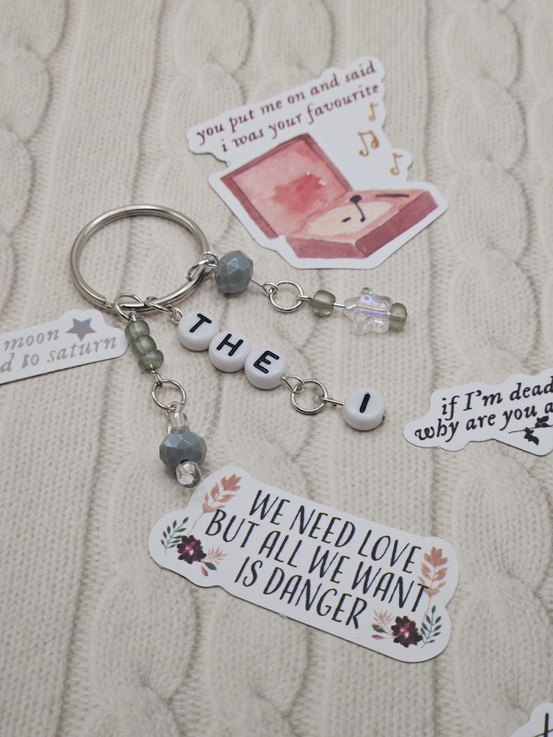May include: A silver keyring with the word "THE I" spelled out in white letter beads. The keyring has decorative grey beads and charms. Stickers with text and illustrations, including "WE NEED LOVE BUT ALL WE WANT IS DANGER", are scattered around.