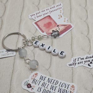 May include: A silver keyring with the word "EXILE" spelled out in white letter beads. The keyring has two grey stone beads and several decorative stickers with song lyrics and illustrations. The background is a white knitted fabric.