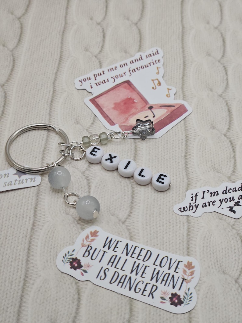 May include: A silver keyring with the word "EXILE" spelled out in white letter beads. The keyring has two grey stone beads and several decorative stickers with song lyrics and illustrations. The background is a white knitted fabric.