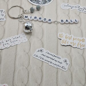 May include: A silver disco ball keyring with the words "MIRROR BALL" spelled out in white letter beads. The keyring includes grey beads and is surrounded by stickers with quotes and a disco ball graphic.