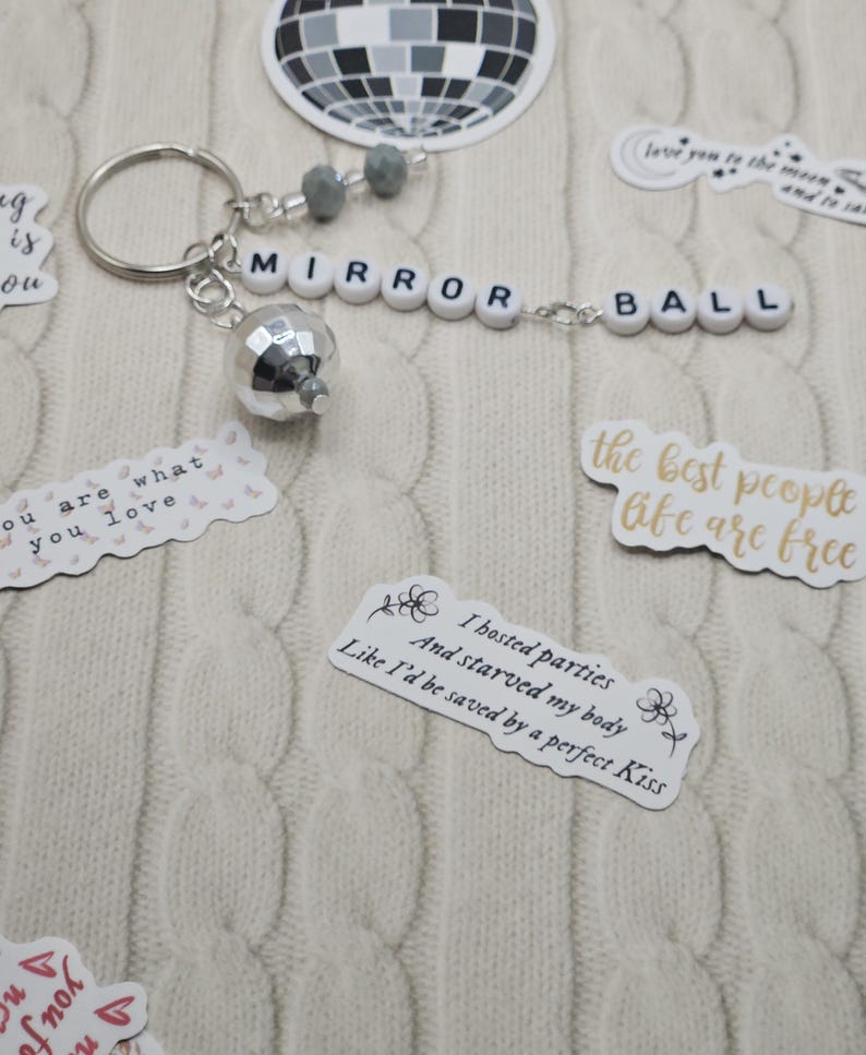 May include: A silver disco ball keyring with the words "MIRROR BALL" spelled out in white letter beads. The keyring includes grey beads and is surrounded by stickers with quotes and a disco ball graphic.