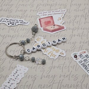 May include: A silver keyring with the word "CARDIGAN" spelled out in white letter beads. The keyring has grey beaded accents and is surrounded by decorative stickers with text and illustrations.