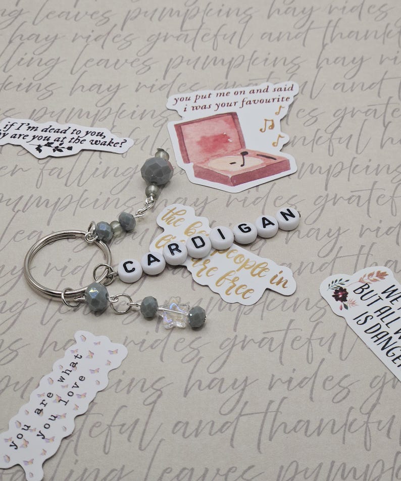 May include: A silver keyring with the word "CARDIGAN" spelled out in white letter beads. The keyring has grey beaded accents and is surrounded by decorative stickers with text and illustrations.