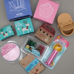 Pode incluir: Uma coleção de brinquedos e acessórios em miniatura, incluindo caixas, pratos e um dispensador PEZ. As caixas apresentam texto e ilustrações, como "WING THINGS" e "BIRCH REINDEER". O dispensador PEZ é rosa e amarelo.