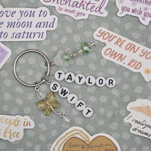 May include: A silver keychain with the name "TAYLOR SWIFT" spelled out in white letter beads. It features a gold butterfly charm and clear bead accents. The background includes stickers with song lyrics and phrases.