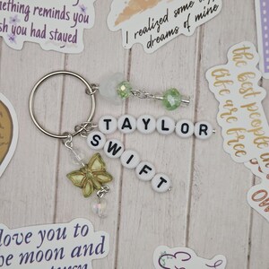 May include: A silver keyring with the name "TAYLOR SWIFT" spelled out in white letter beads. A gold butterfly charm and green beads dangle from the keyring. The background includes stickers with inspirational quotes.