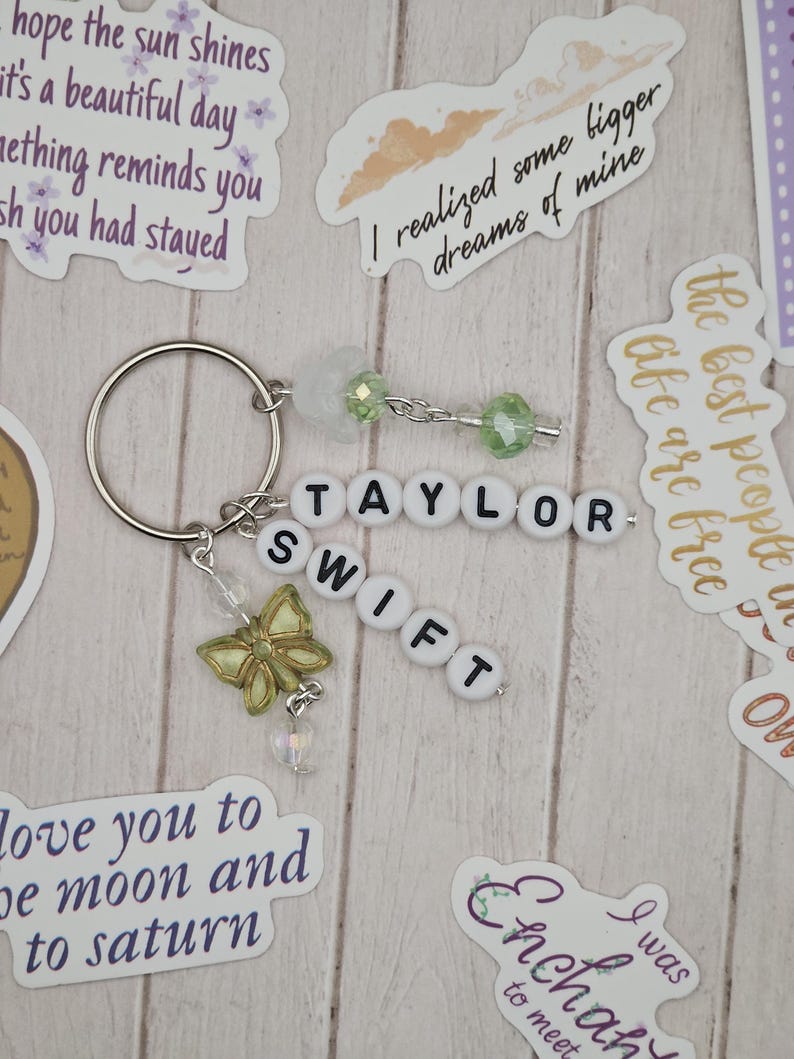 May include: A silver keyring with the name "TAYLOR SWIFT" spelled out in white letter beads. A gold butterfly charm and green beads dangle from the keyring. The background includes stickers with inspirational quotes.