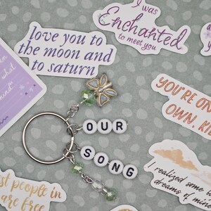 May include: A silver keychain with the words "OUR SONG" spelled out in white letter beads. It features a butterfly charm and green beaded accents. Surrounding the keychain are various stickers with phrases like "love you to the moon and to saturn".