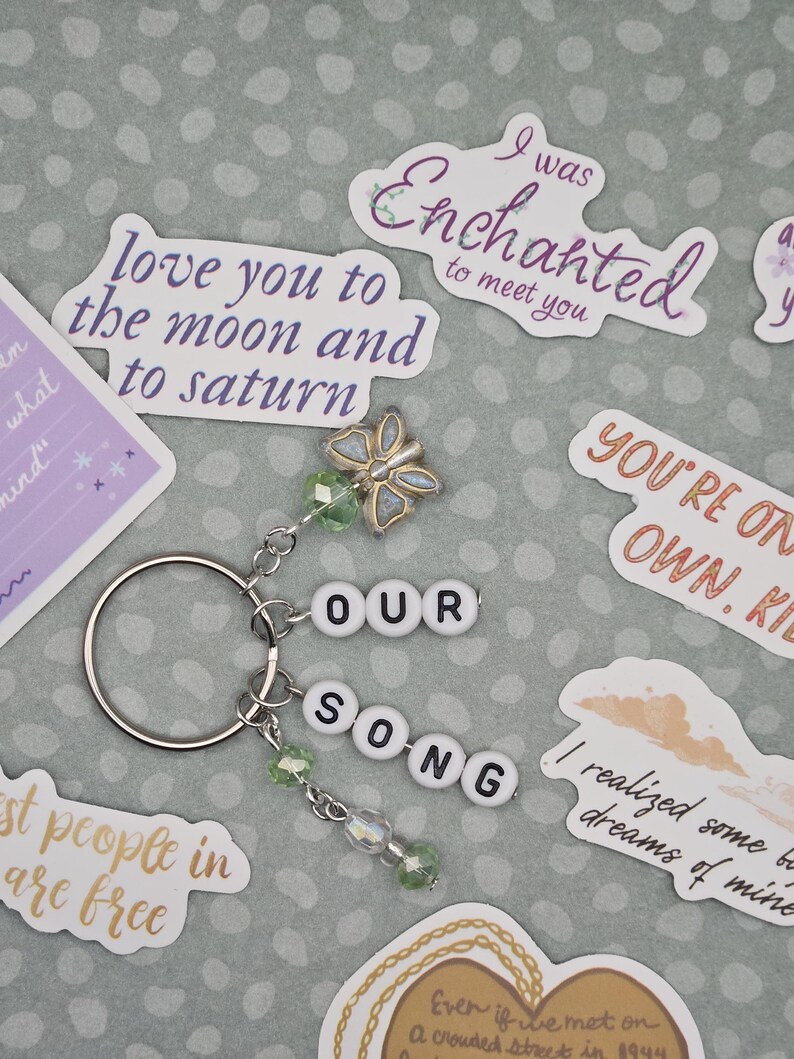 May include: A silver keychain with the words "OUR SONG" spelled out in white letter beads. It features a butterfly charm and green beaded accents. Surrounding the keychain are various stickers with phrases like "love you to the moon and to saturn".